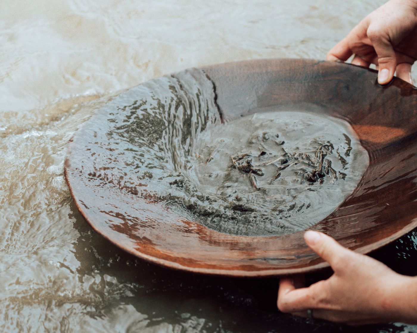 Unas manos sostienen la batea de madera sobre el agua del río Cauca, con la arena donde se busca el oro