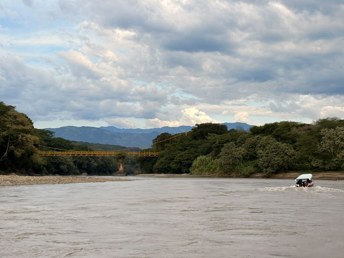 La lancha con pasajeros cruzando el río Cauca, pequeña entre los árboles de la orilla