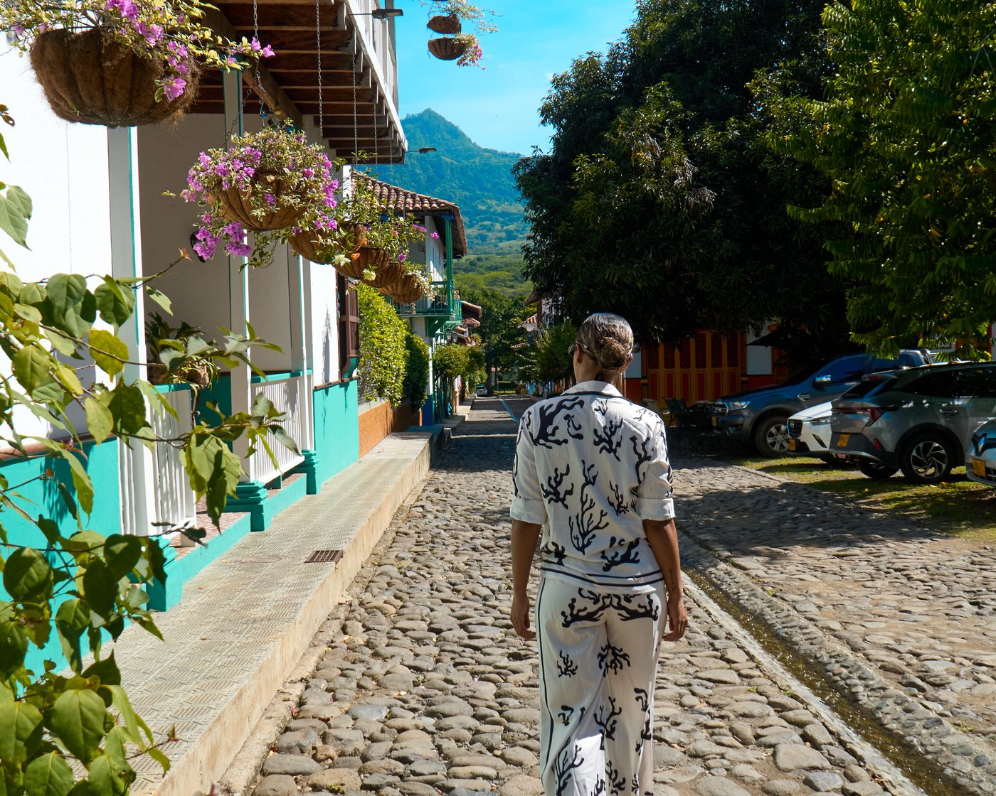 Una persona camina por la calle empedrada entre casas con materas de flores colgadas, con las montañas al fondo